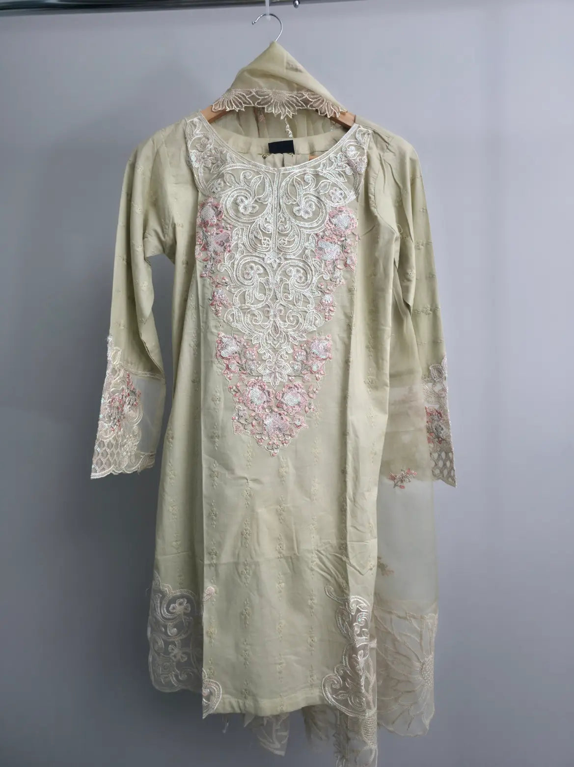 A light beige traditional tunic dress with intricate white and pink floral embroidery, hanging on a wooden hanger against a plain gray wall.
