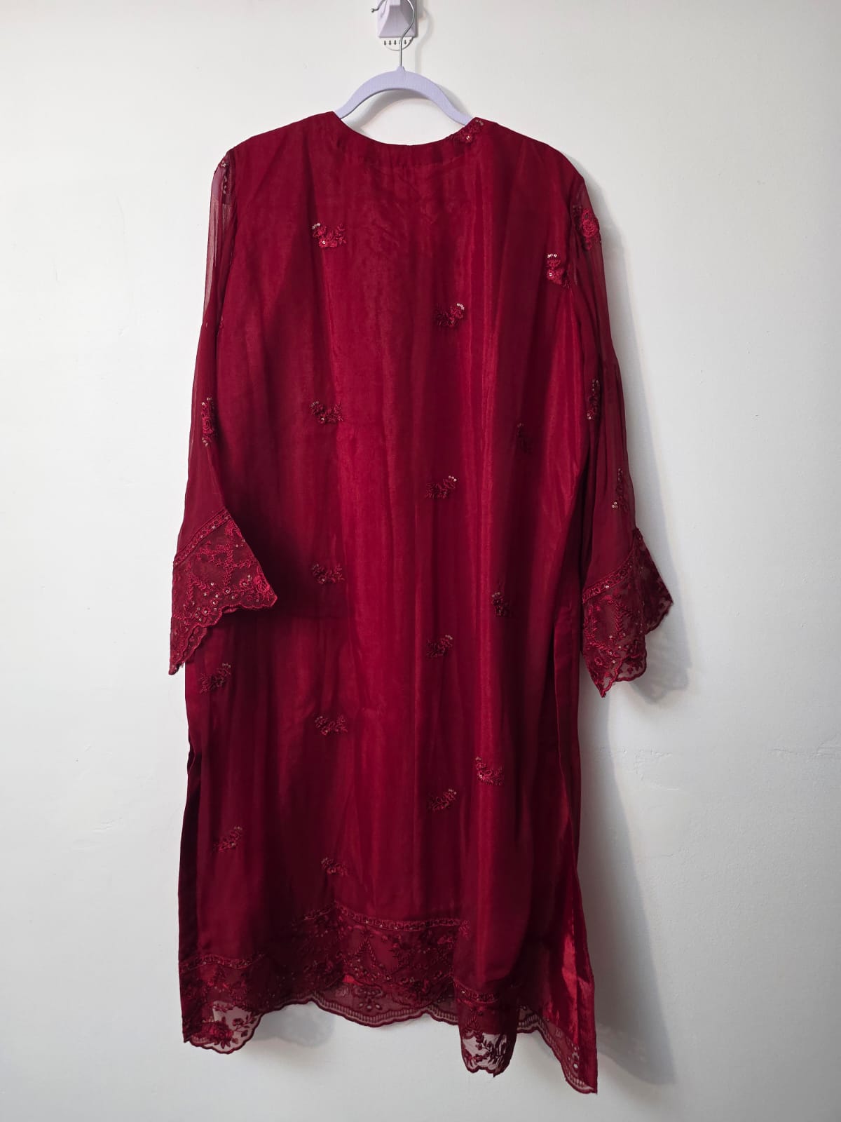 A deep red sheer tunic dress with floral embroidery and lace trim, hanging on a white plastic hanger against a plain off-white wall.