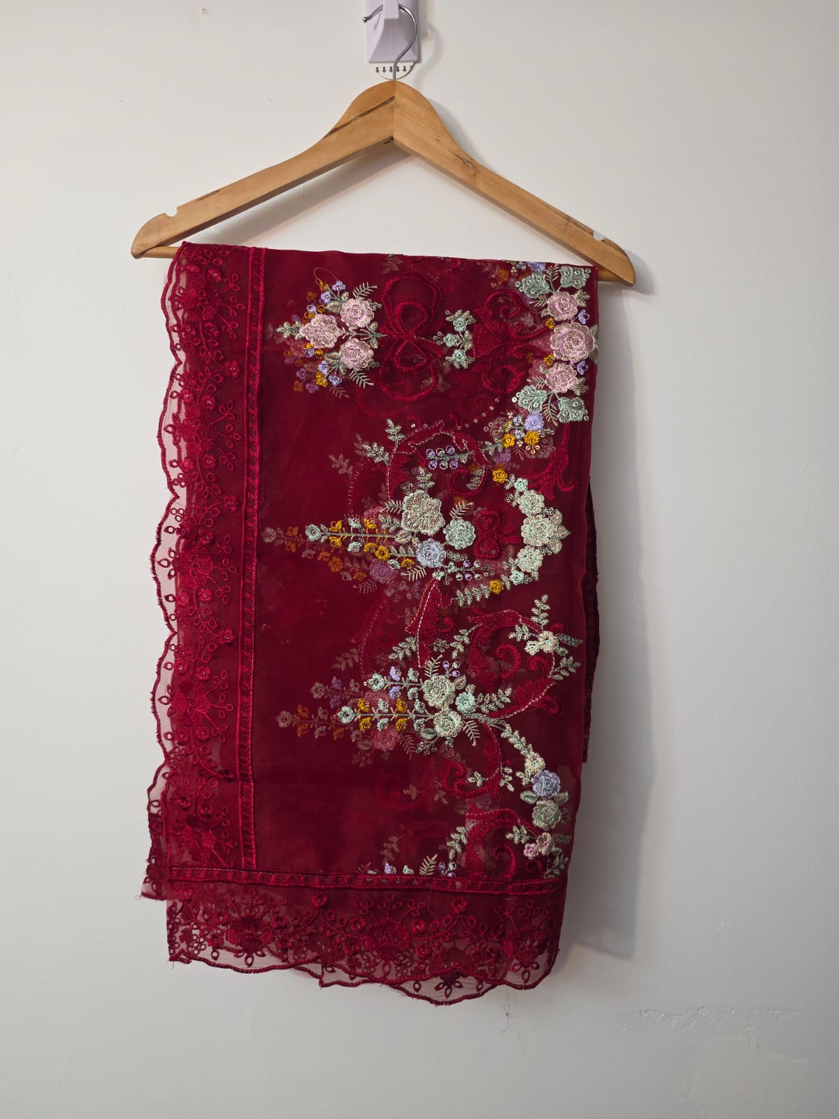 A deep red embroidered lace fabric, likely a sash, scarf, or decorative textile, hanging on a wooden hanger against a plain off-white wall. The fabric features intricate floral embroidery in shades of pink, white, yellow, and green, with matching red lace trim along the edges.