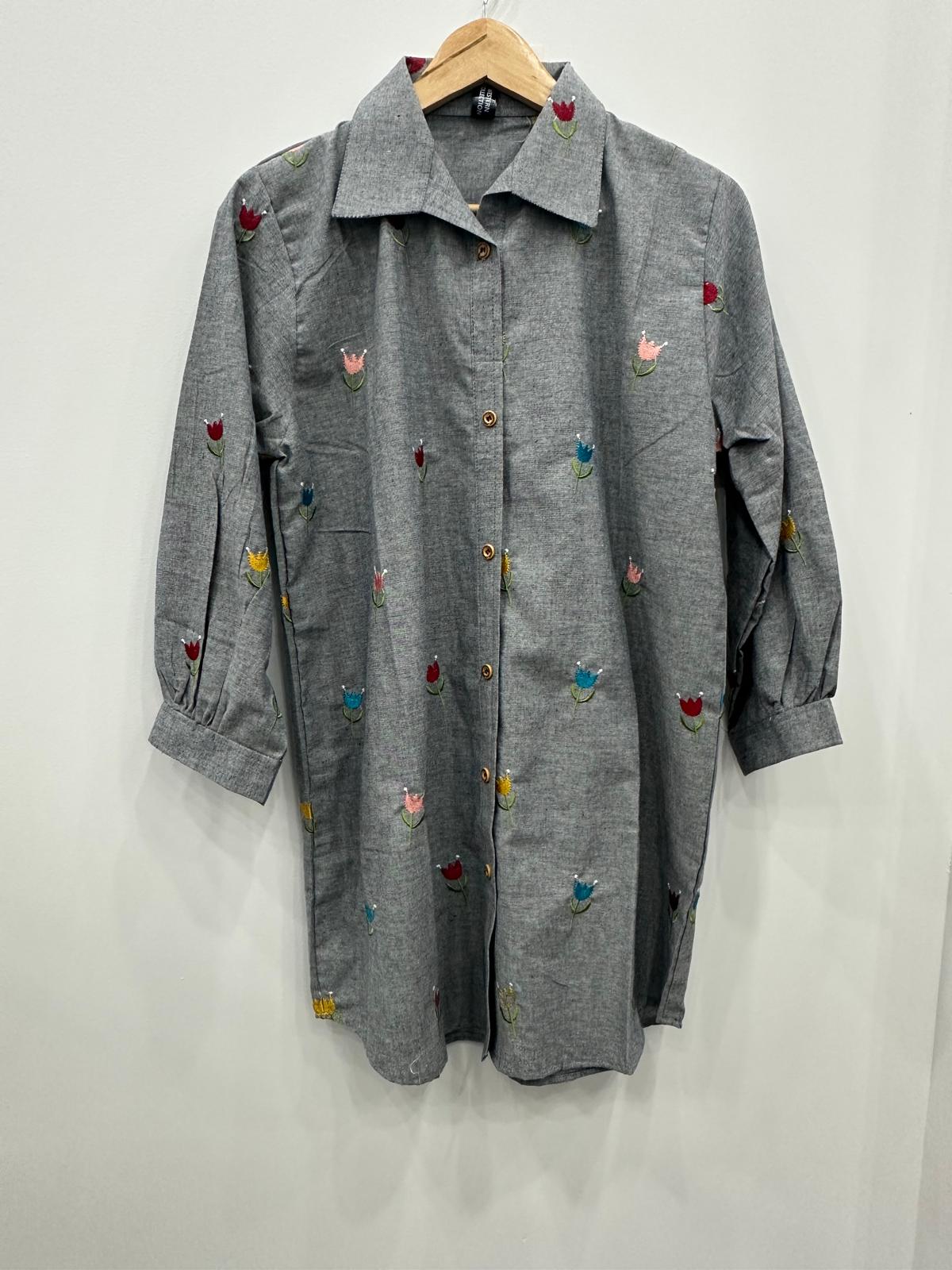 A gray long-sleeve button-up shirt with small colorful embroidered floral motifs, hanging on a wooden hanger against a plain light gray wall.