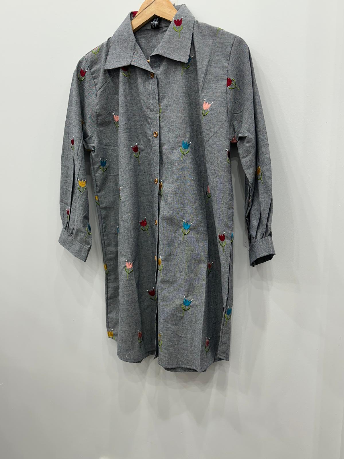 A gray long-sleeve button-up shirt dress with small colorful embroidered floral motifs, hanging on a wooden hanger against a plain white wall.