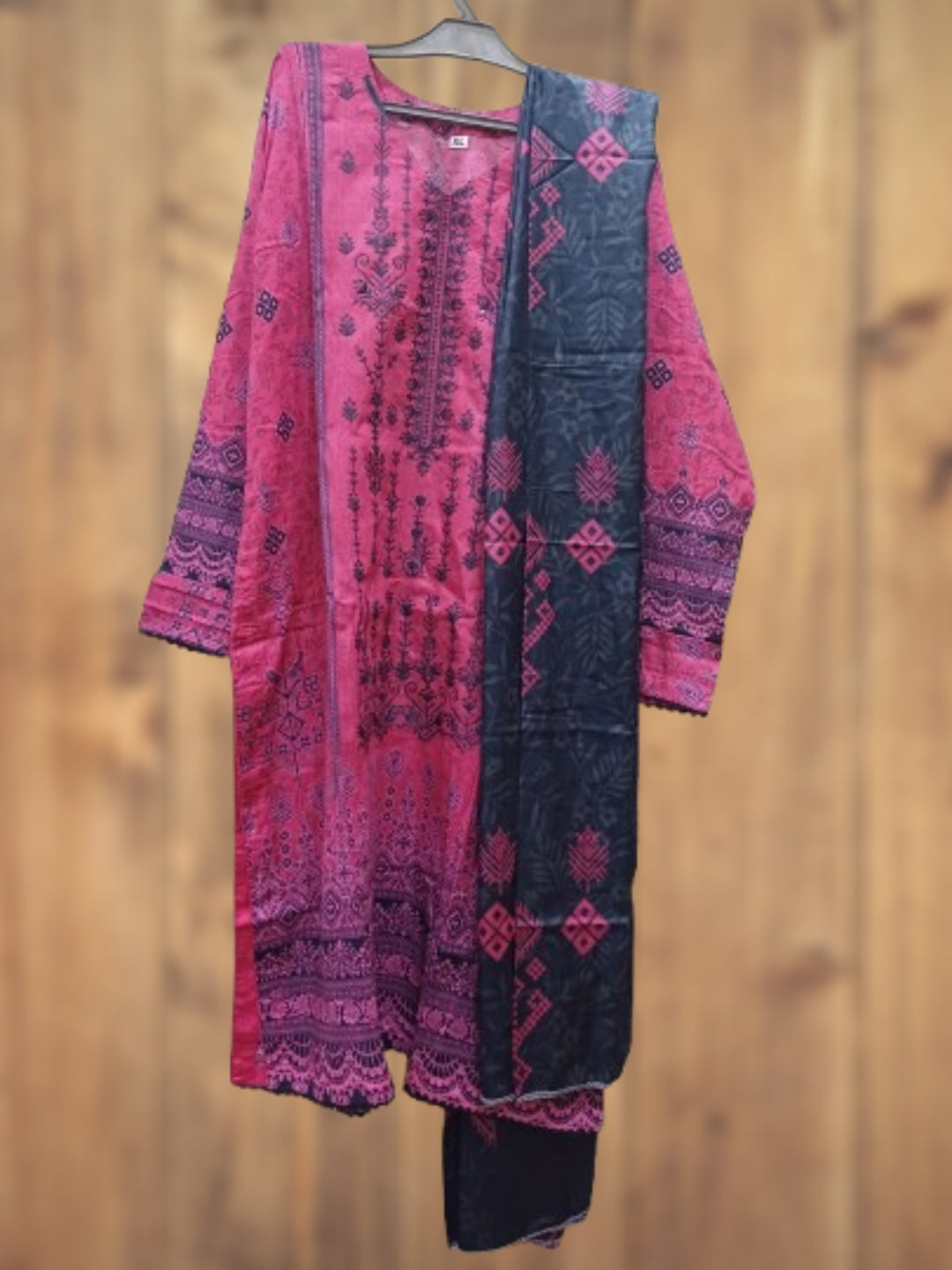A three-piece traditional Indian salwar suit set featuring a pink and black printed kurta, matching trousers, and a coordinating dupatta, displayed on a hanger against a wooden wall background.