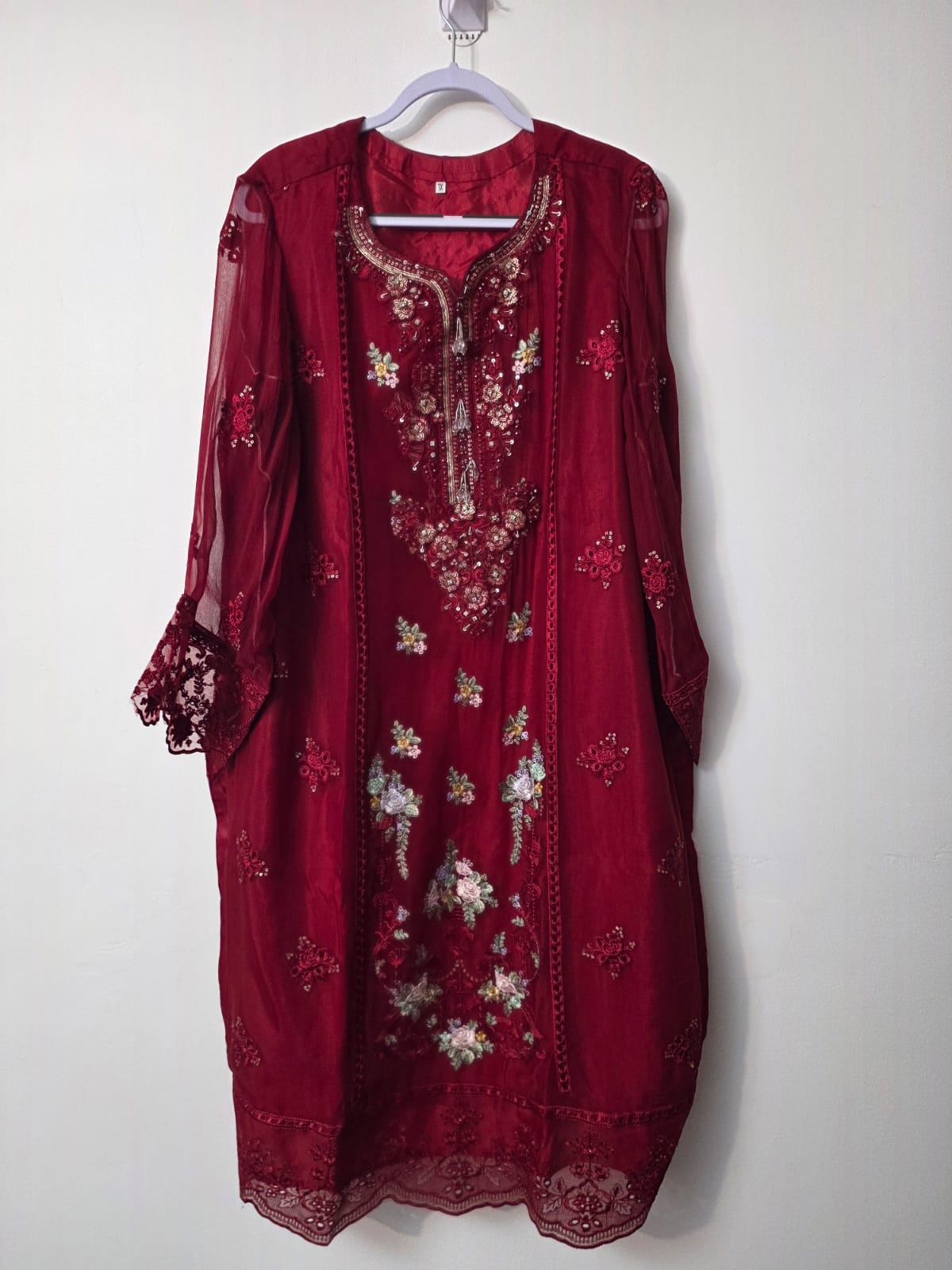 A deep red embroidered kurta with sheer sleeves and floral embroidery, hanging on a white hanger against a plain off-white wall.
